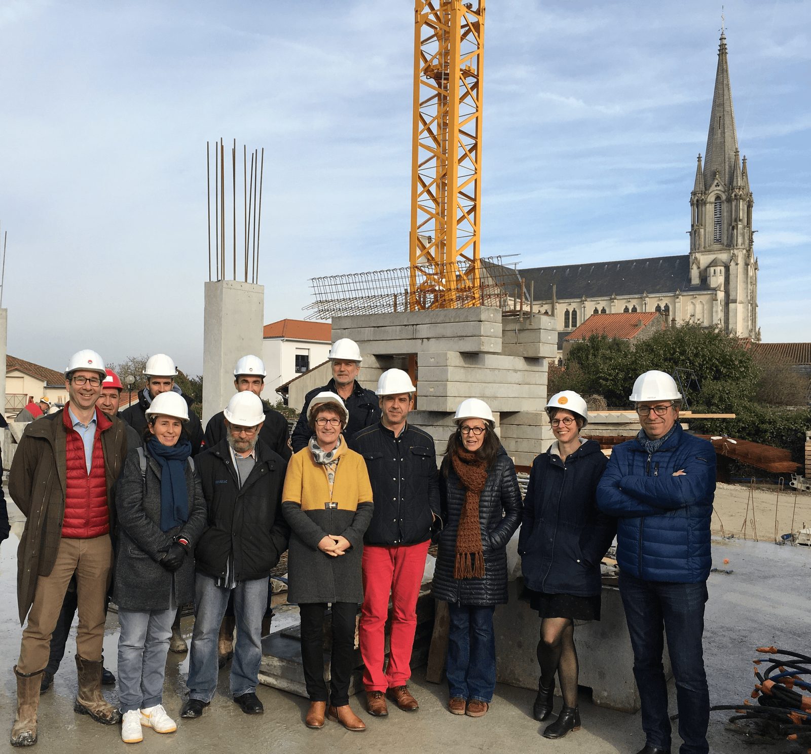 Visite de chantier en tenue de sécurité : intervenants réunis devant les poteaux en béton et la grue, église en arrière-plan
